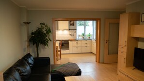 A living room with a black leather sofa, a wooden coffee table, and a TV stand. A large plant is in the corner, and there is a kitchen in the background with white cabinets and a window.