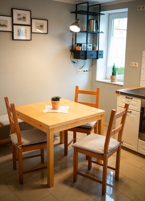 Ein Essbereich mit einem Holztisch und Stühlen, einem kleinen Topfpflanzen und einem an der Wand befestigten Regal mit gerahmten Bildern.
