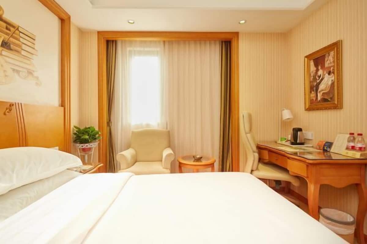 A hotel room with a bed, two chairs, a desk, a small table, a plant, a painting, and a window with curtains.
