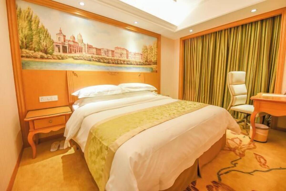 A hotel room with a bed, a nightstand, a desk, a chair, and a painting on the wall.