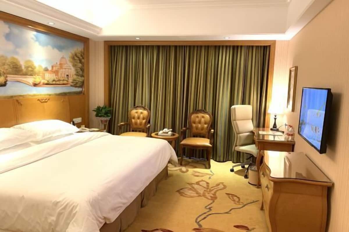 A hotel room with a large bed, two armchairs, a desk with a TV, and a painting on the wall.