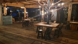 Ett utomhusområde med plats för måltider, utrustat med träbord och stolar, en central, trädlika konstruktion och en eldstad.