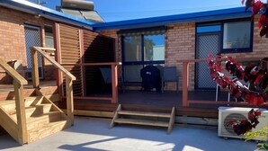 Un porche cubierto con una escalera de madera que lleva a una terraza, una parrilla y una zona de estar con sillas.