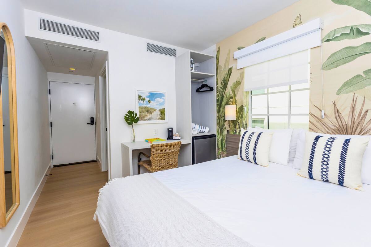 A hotel room with a bed, a desk, a wardrobe, and a window with a view of palm trees.