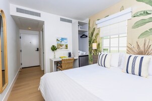 Una habitación de hotel con una cama grande, un escritorio con silla, un armario y una ventana con vistas a palmeras.