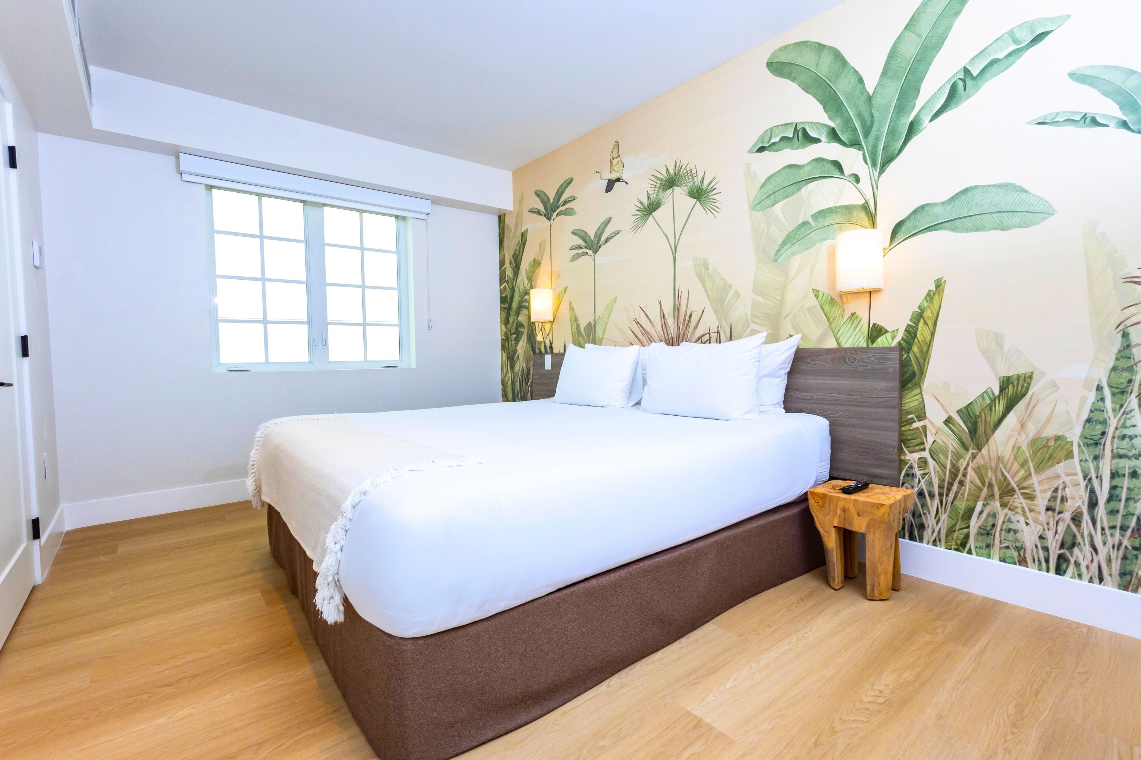 A bedroom with a bed, a window, a wall with a plant mural, and a nightstand.