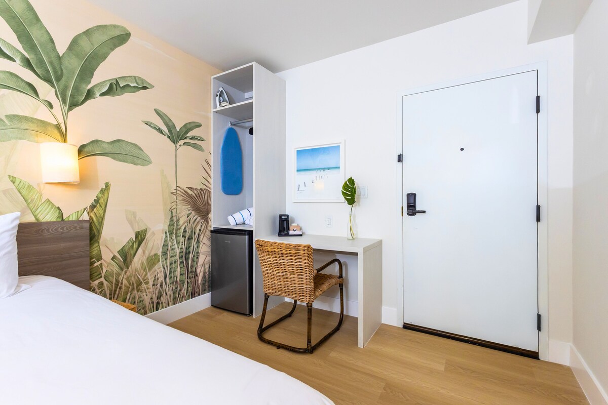 A hotel room with a bed, a desk with a chair, a wardrobe, a refrigerator, and a large potted plant.
