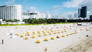 Una playa con numerosos sombrillas amarillas y blancas y sillones de playa, arena y un paisaje urbano con edificios altos al fondo.