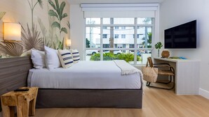 A modern hotel room with a large bed, a desk with a TV, a window with a city view, and a plant. - Casa Ocean (Miami Beach)