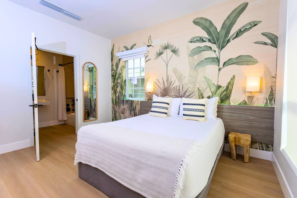 A bedroom with a large bed, a wooden nightstand, a mirror, and a tropical-themed wall mural.