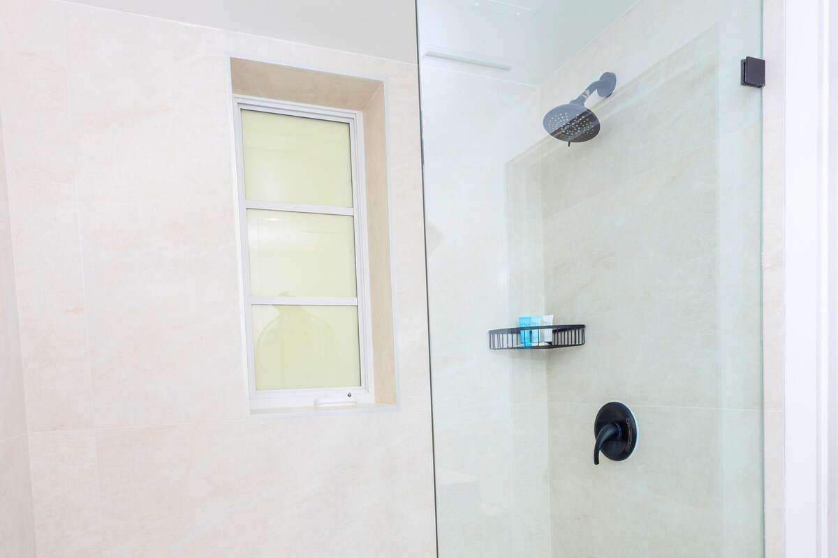 A glass-enclosed shower with a handheld showerhead, a small shelf with toiletries, and a window.