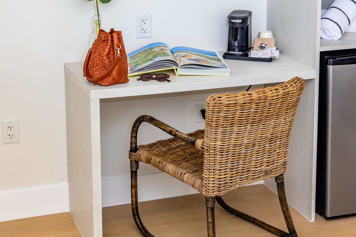 A white desk with a wicker chair, a plant, a coffee maker, and a framed picture of a beach.