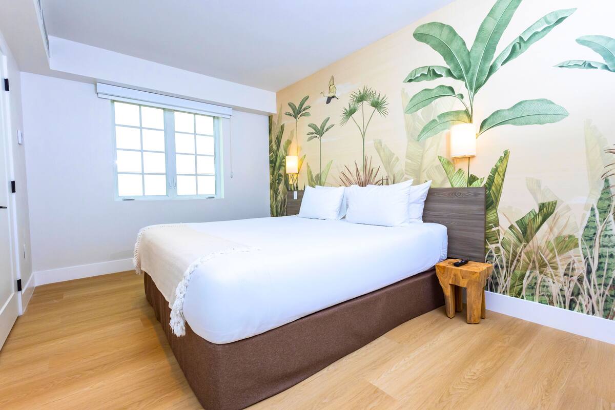 A bedroom with a large bed, a wooden nightstand, a window, and a wall mural of tropical plants.