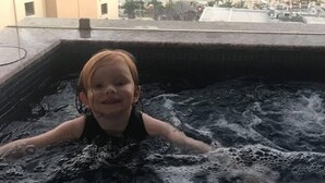 Criança em banheira de hidromassagem, com vista para uma cidade costeira e edifícios ao fundo.