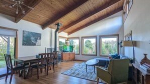 A spacious living room with wooden floors, a dining area with a table and chairs, a television, and large windows offering a view of the outdoors.