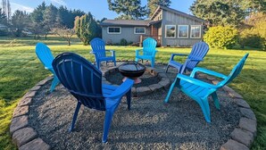 A backyard with a fire pit surrounded by blue Adirondack chairs, a wooden deck, and a house in the background.