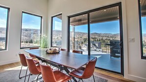 A spacious dining area with a large table and chairs, large windows offering a view of a town and hills, and a modern architectural design.