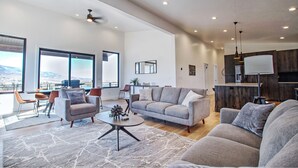 A spacious living area with a modern sofa set, a coffee table, and a dining area with chairs. There is a kitchen visible in the background with wooden cabinets and a hanging light fixture.