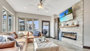 A spacious living room with a leather sofa, a tufted ottoman, a fireplace, and a large TV mounted on a stone wall.