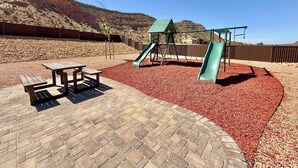 A playground with a slide and swings, surrounded by a brick patio and mulch. - Maison Sans Souci - Kanab Home, Pool & Views (Kanab)