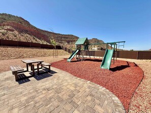Ein Spielplatz mit Rutsche und Schaukeln, umgeben von einer Pflasterterrasse und Rindenmulch.
