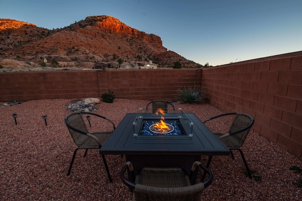 Un patio al aire libre con una mesa y sillas alrededor de un hogar, rodeado por un muro de ladrillo y piedras pequeñas.