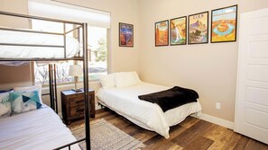 A bedroom with a bunk bed, a single bed, a nightstand, a lamp, and a rug. There are four framed posters on the wall.