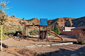 Un parque infantil con un tobogán y columpios, rodeado de un paisaje montañoso y un edificio al fondo.