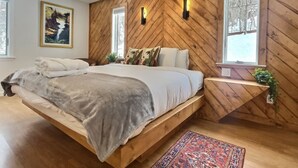 Une chambre avec un mur en bois à motifs en chevron, un lit recouvert d’une couverture grise, deux lampes fixées au mur, une fenêtre donnant sur des arbres et une petite étagère avec une plante.