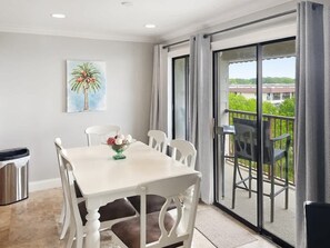A dining area with a white table and chairs, a trash bin, and a painting of a palm tree on the wall. - Beachfront Hilton Head Resort 2BR (AR-412): Tennis, Pickleball & Largest Pool (Hilton Head Island)