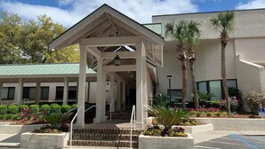 A building with a covered entrance, a parking lot, and a landscaped area with palm trees and flowers. - Beachfront Hilton Head Resort 2BR (AR-412): Tennis, Pickleball & Largest Pool (Hilton Head Island)