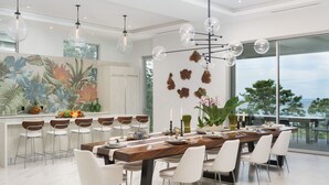 A modern dining area with a long wooden table, white chairs, and a large window offering a view of the outdoors.