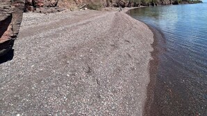 Una playa de guijarros con un acantilado rocoso y árboles, con vistas a una masa de agua tranquila.