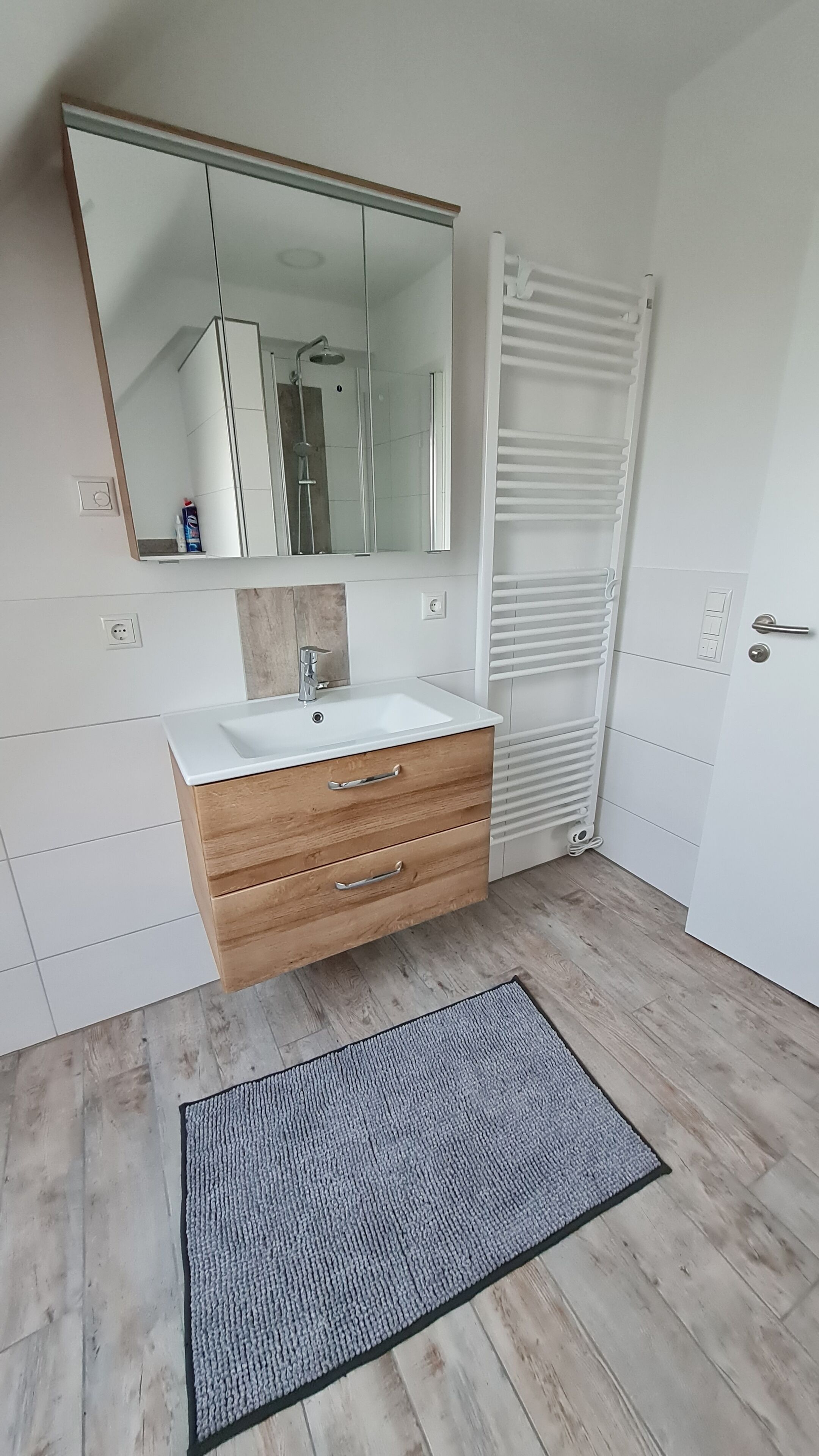 A bathroom with a white sink, wooden vanity, a large mirror, a towel rack, and a mat on the floor.