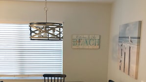 A dining area with a round table and six chairs, a hanging light fixture, a 'Welcome to the Beach' sign, and a seascape painting on the wall. - Sunsets at Gulf of Mexico (Venice)
