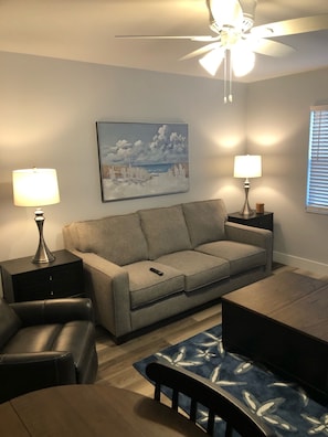 A living room with a gray sofa, two side tables with lamps, a ceiling fan, a painting of a beach scene, and a wooden coffee table.