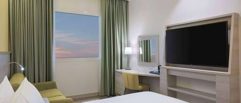 Habitación de hotel con cama, escritorio, silla, televisor y ventana con cortinas.