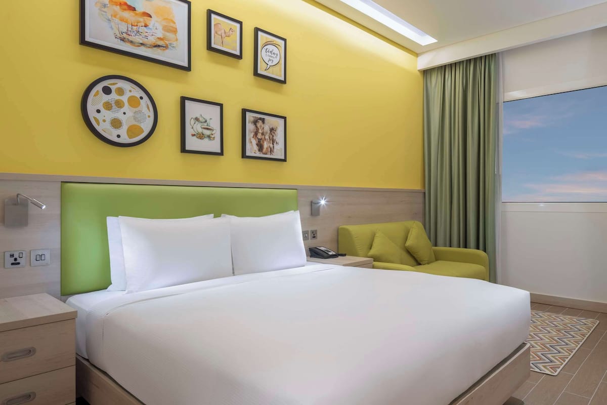 A hotel room with a large bed, a green headboard, a yellow accent wall, and artwork.