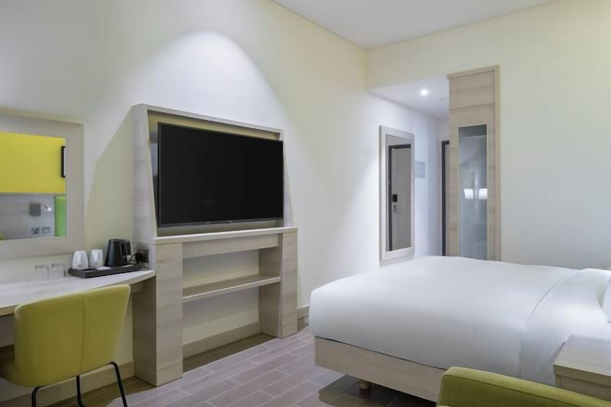 Habitación de hotel con cama, escritorio, silla, televisor y espejo.