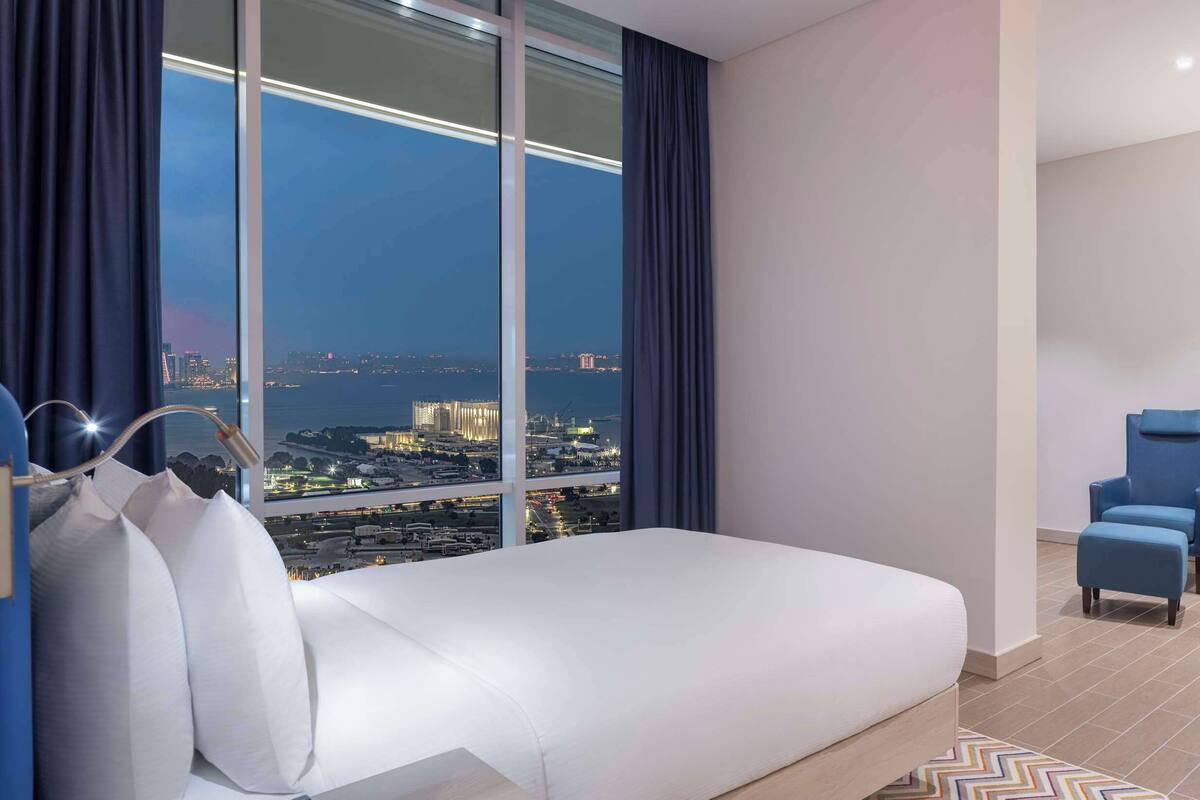 A hotel room with a bed, a desk, a chair, a TV, and a large window with a city view.