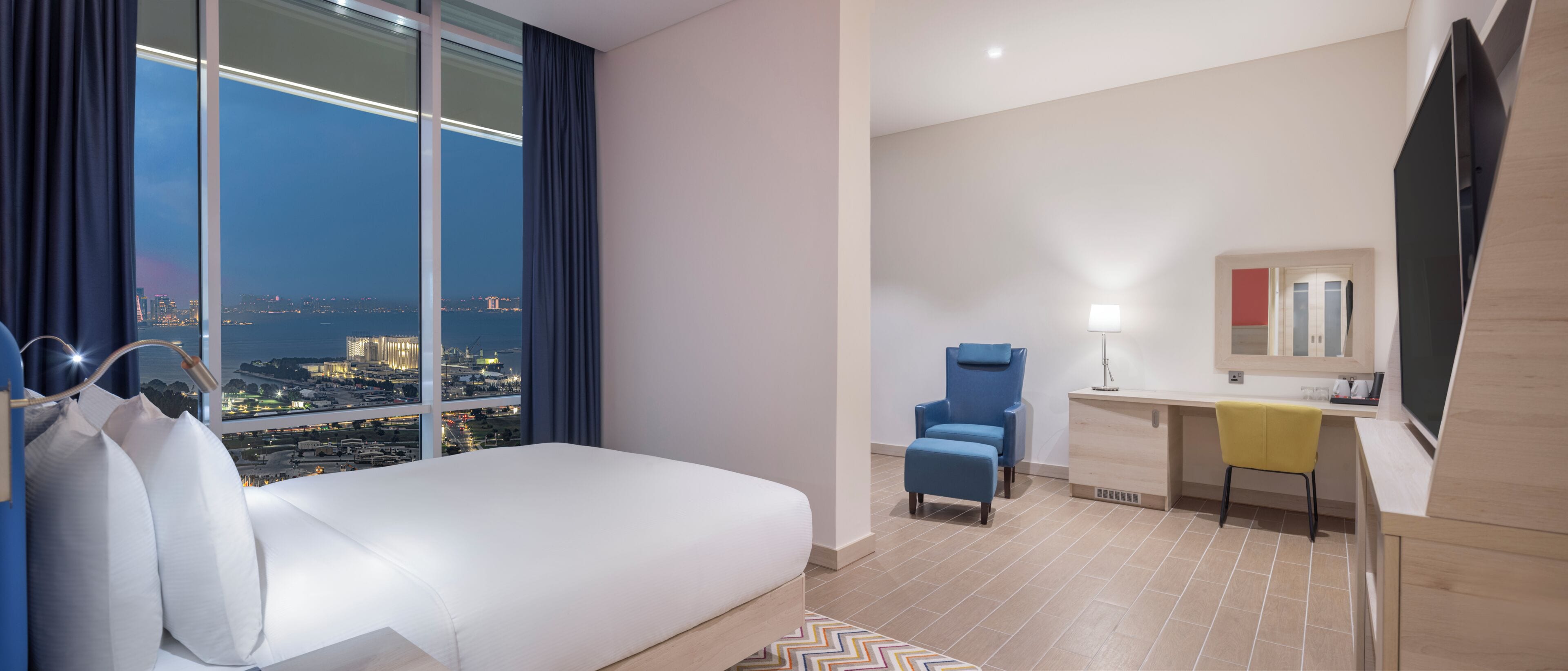 A hotel room with a bed, a desk, a chair, a mirror, and a view of a cityscape.
