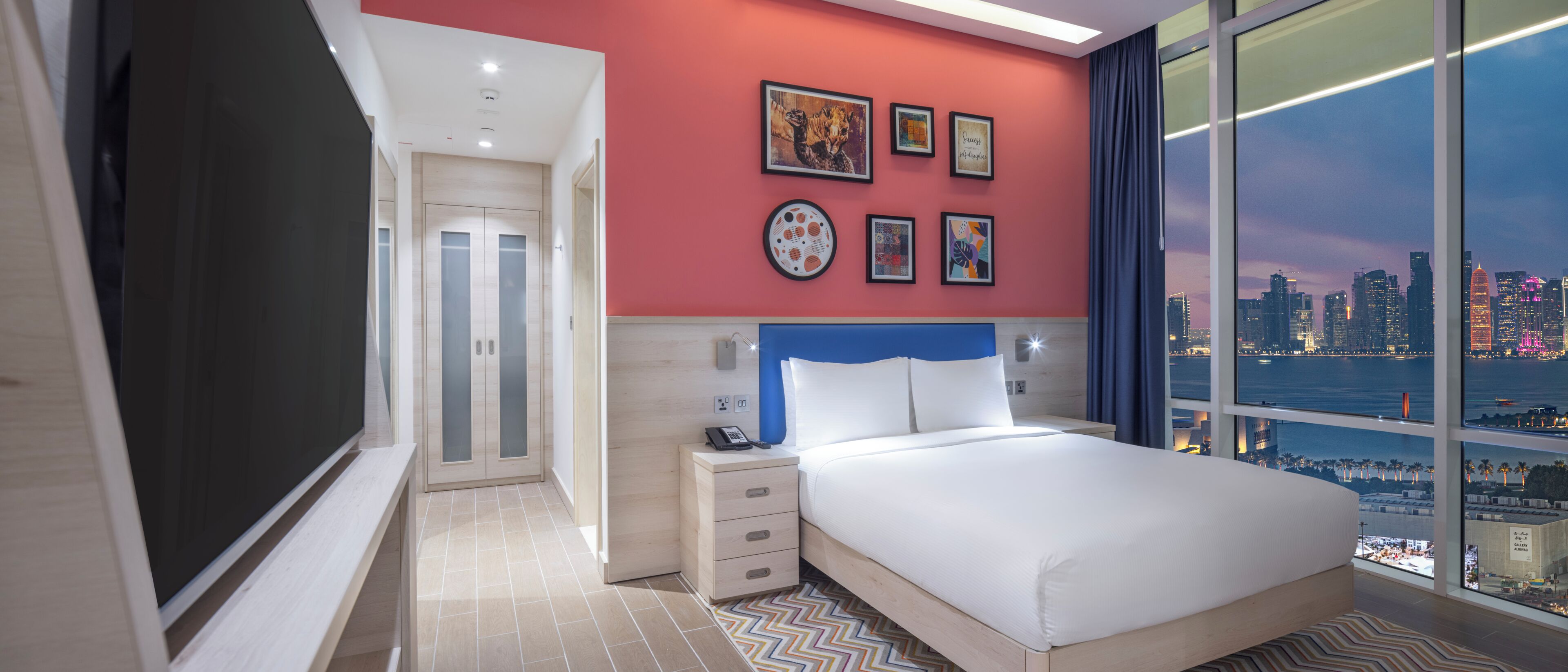 A hotel room with a bed, a nightstand, a wall with framed artwork, and a large window offering a city view.