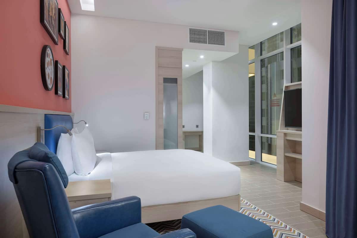A modern hotel room with a large bed, a blue chair, a desk, and a wardrobe.