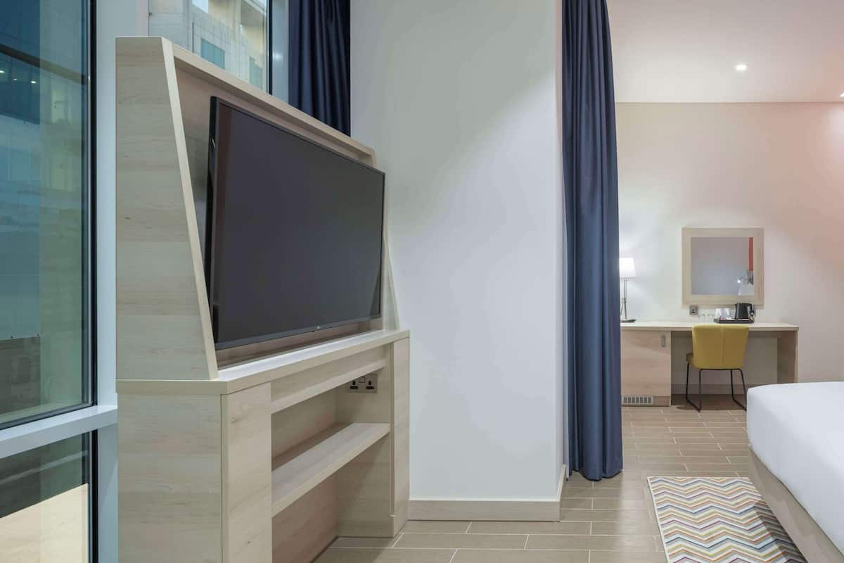 A modern hotel room with a large flat-screen TV, a desk with a chair, a bed, and a bathroom visible through an open door.
