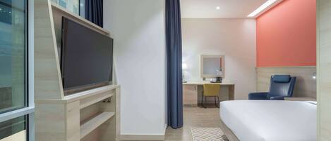 A modern hotel room with a large flat-screen TV, a desk with a chair, a bed, and a bathroom visible through an open door.