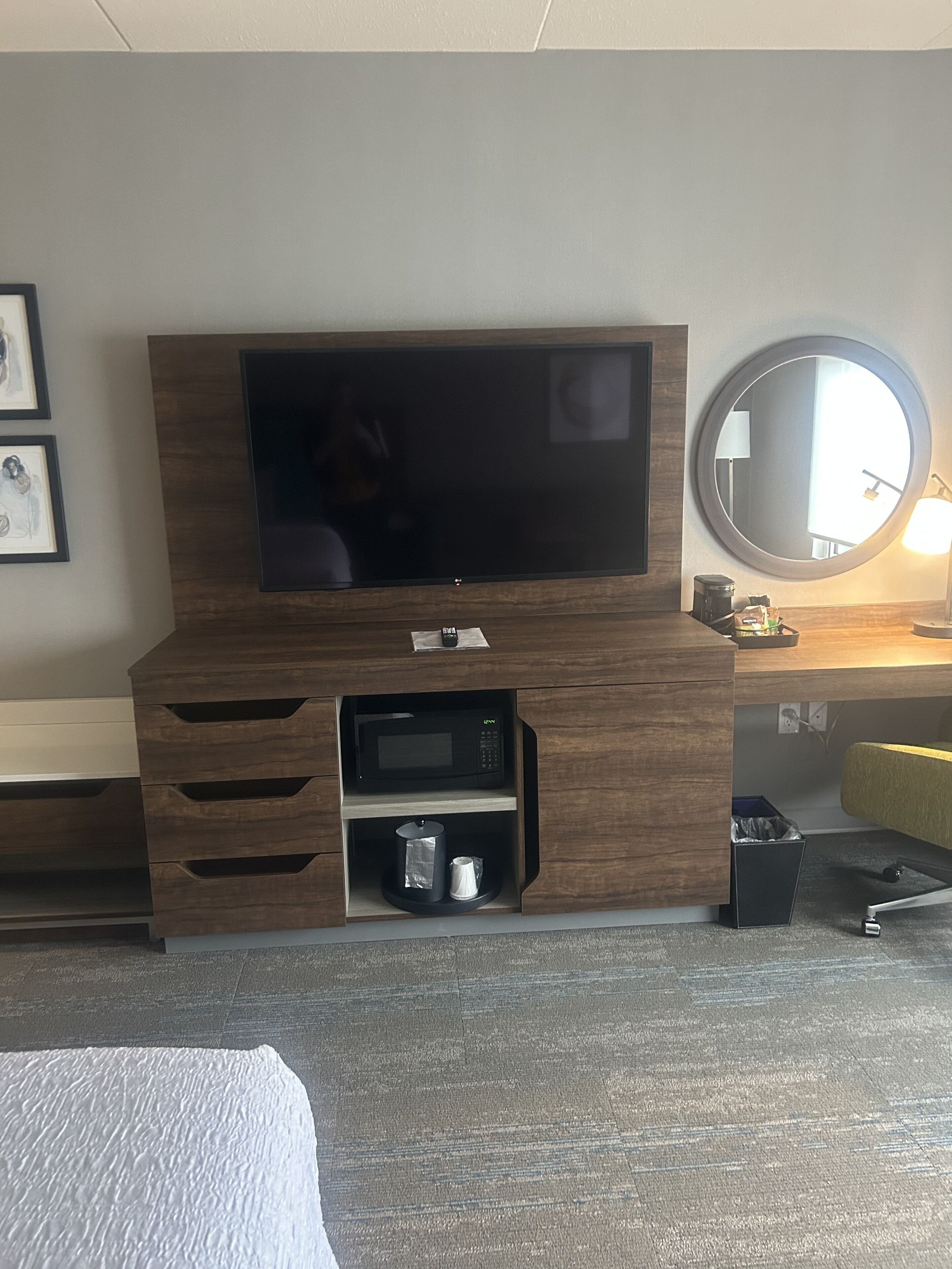 Habitación de hotel con un televisor de pantalla plana grande montado en un mueble de madera, una cafetera pequeña, un cesto de basura y un espejo redondo en la pared.