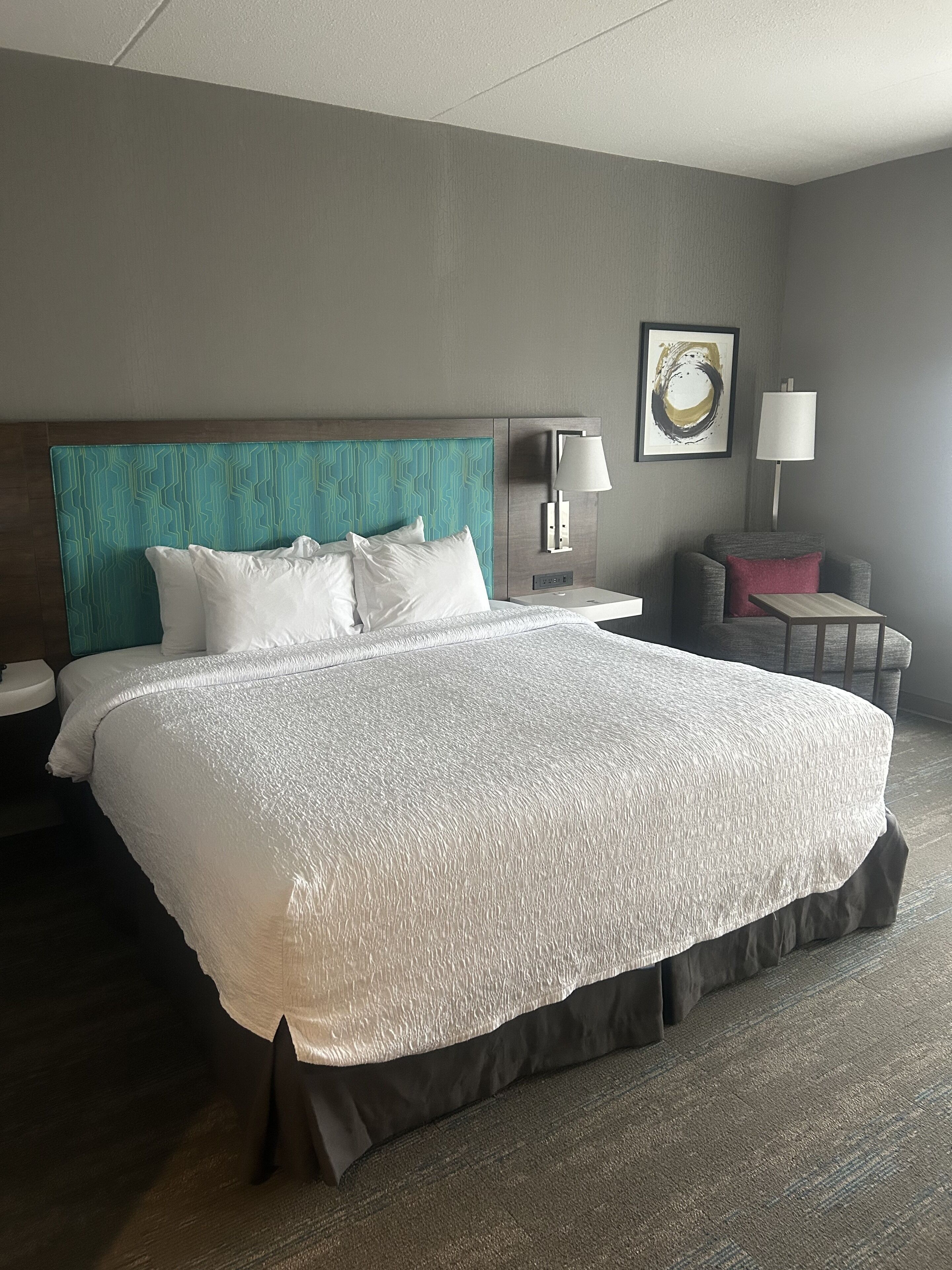 A hotel room with a large bed, a nightstand, a chair, and a wall-mounted lamp.