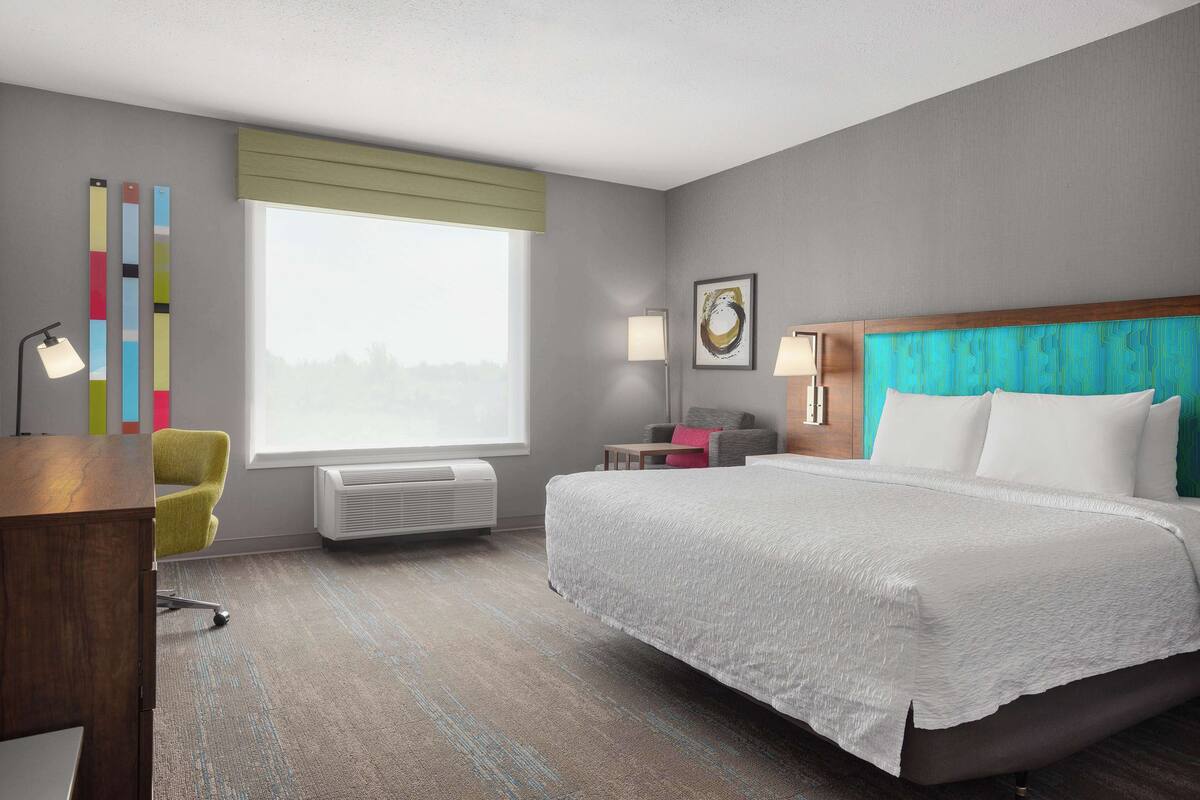 Habitación de hotel con una cama grande, un escritorio, una silla, una ventana con persianas, aire acondicionado de pared y un cuadro enmarcado.