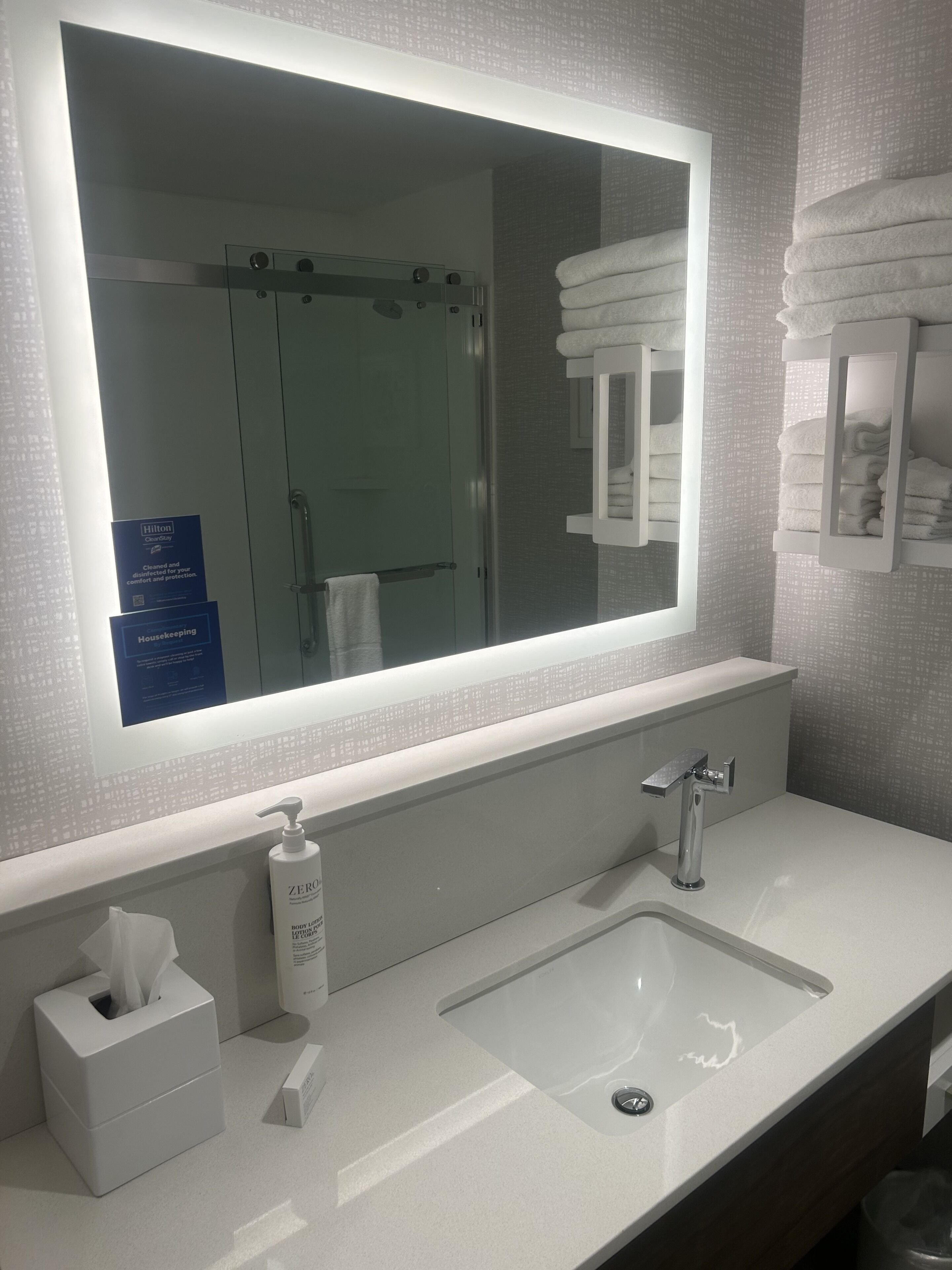 Un baño moderno con un espejo grande, un lavabo, un perchero para toallas y un dispensador de gel antibacterial.
