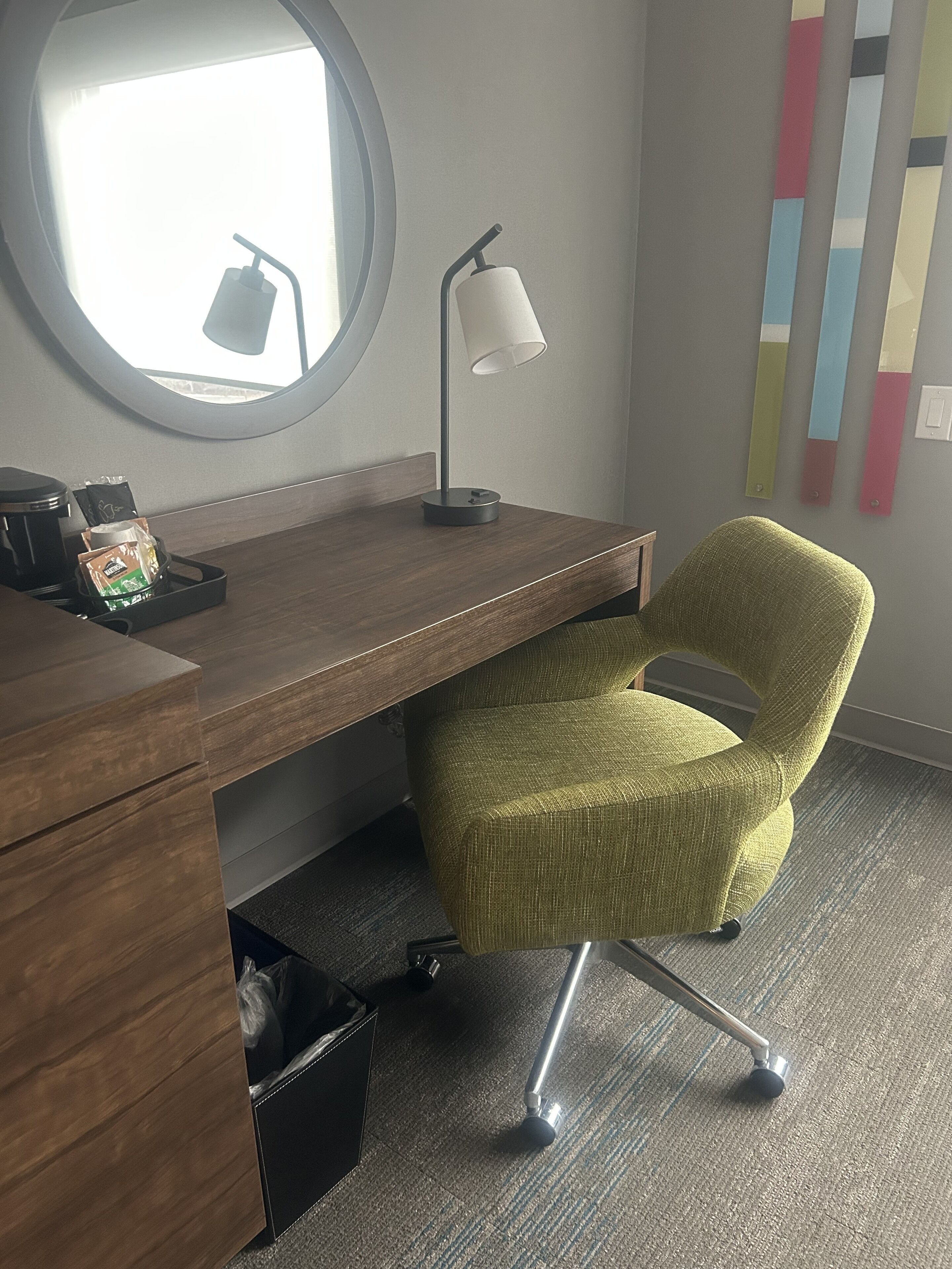 A wooden desk with a lamp, a green chair, a trash bin, and a coffee maker.